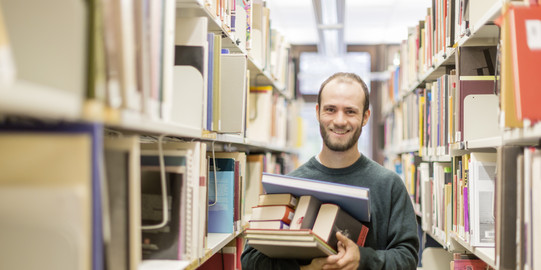 A student standing between two bookshelves is holding a pile of books.