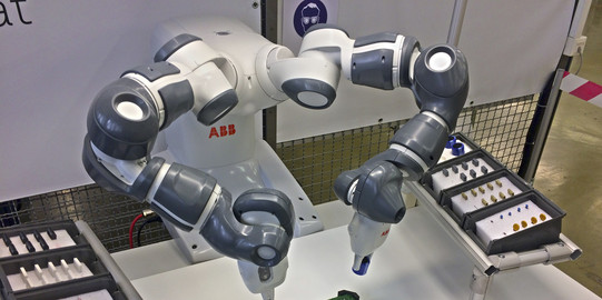 Zweiarmiger Roboter platziert THT-Bauteile auf Platine. Auf beiden Seiten liegen weitere THT-Bauelemente vereinzelt bereit.